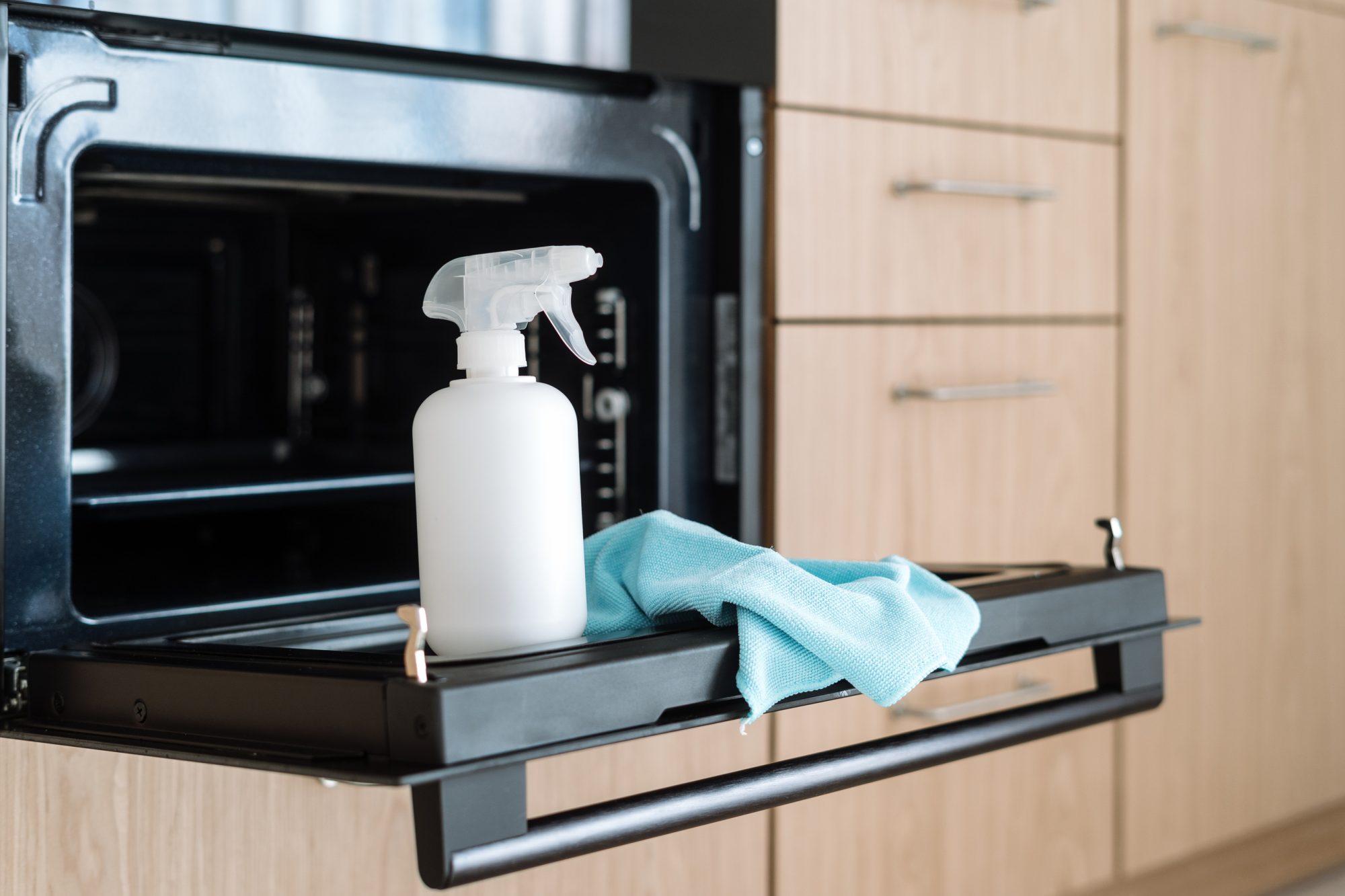 White spray bottle with detergent and cloth standing on open oven door. Advertising concept of cleaning household appliance at home kitchen